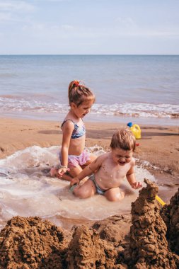 Küçük kızlar dalgalarda neşeyle oynarlar, deniz köpüğünde gülerek ve su sıçratarak. Deniz kenarında eğlenceli bir yaz anı.