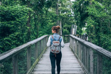 A young woman with a backpack walks alone on a wooden bridge surrounded by green forest. Peaceful travel and connection with nature.