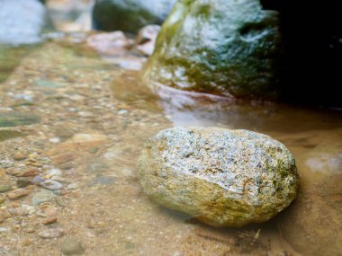 Görünür kum ve çakıl taşlarıyla sığ, şeffaf bir dere suyuna batırılmış granit bir kayanın yakın çekimi. Arka planlar, toprak tonlama tasarımları ve çevresel içerik için mükemmel
