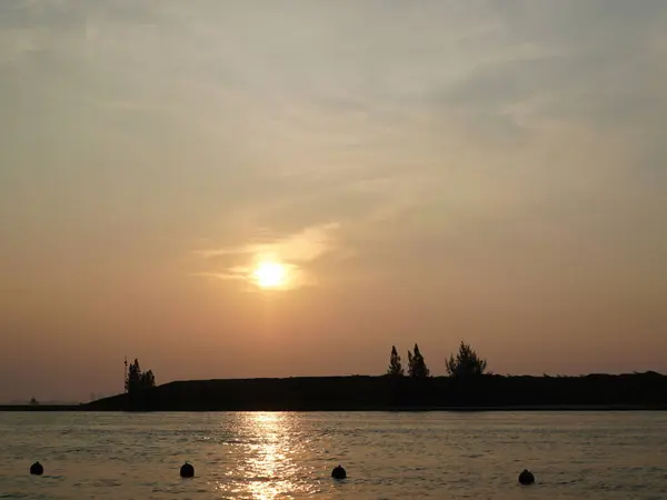 Durgun suların üzerinde batan güneşin sıcak tonlarını yakalayan altın saat sahnesi. Nazik yansıma, ufuktaki yumuşak bulutlar ve karanlık ağaç siluetleriyle güçlendirilmiş sakin bir ruh hali yaratır.