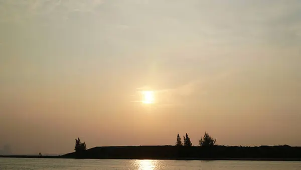 Minimalist günbatımı fotoğrafı ince bulut desenleri, parlayan bir güneş ve alttaki sakin sularla geniş bir gökyüzünü gösteriyor. Sahne, rahatlama, dinginlik ve doğal güzelliği çağrıştırıyor.