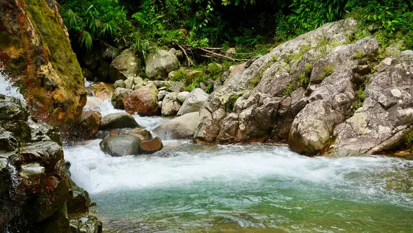Zümrüt yeşili su, vahşi bitki örtüsü ve şelale ile çevrili kırmızı tonlu bir kayanın altında hızla akıyor. Çevresel seyahat ve doğa kaçışı görselleri için mükemmel.