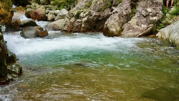 Zümrüt yeşili su, vahşi bitki örtüsü ve şelale ile çevrili kırmızı tonlu bir kayanın altında hızla akıyor. Çevresel seyahat ve doğa kaçışı görselleri için mükemmel.