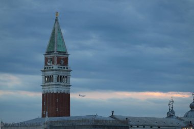 Il campanile di San Marco svetta tra le nuvole mentre un aereo attraversa il cielo. Bir uçak gökyüzünü geçerken St. Marks çan kulesi bulutların arasından yükseliyor..
