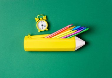 Renkli kalemler ve yeşil zemin üzerindeki saat ile sarı kalem şeklinde bir çanta..