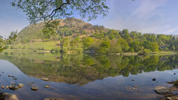 Berrak mavi gökyüzünün altındaki yemyeşil tepeleri ve ağaçları yansıtan sakin bir göl manzarası..