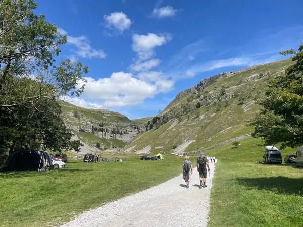 Malham, iki yürüyüşçü yemyeşil bir vadide çakıllı bir patika boyunca yürüyor. Yuvarlanan tepeler ve uçsuz bucaksız gökyüzü manzaralı bir manzara yaratır. Çadırlar ve arabalar da mevcut..