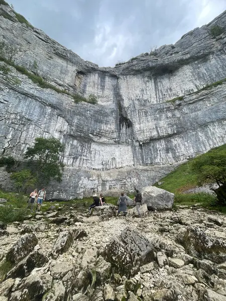 Malham, insanlar bulutlu bir gökyüzünün altındaki yemyeşil yemyeşil kayalıkların yanındaki kayalık arazide yürüyüş yapıyorlar..