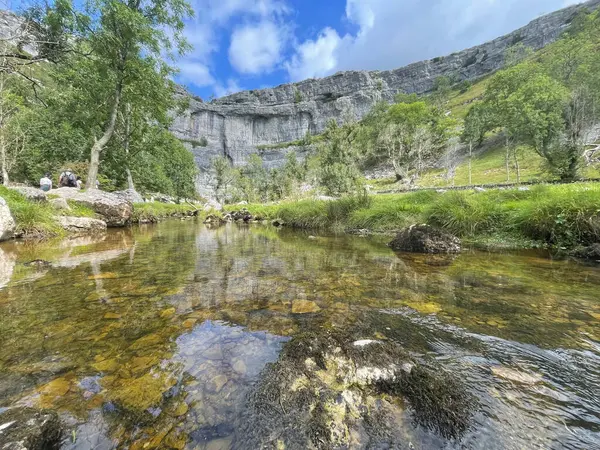 Malham, berrak bir nehir, parlak mavi gökyüzünün altında yemyeşil bir çerçeve ile çevrelenmiş engebeli bir yamaç yüzünü yansıtıyor..