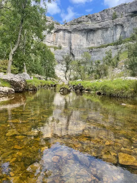 Malham, bulutlu bir gökyüzünün altında, ağaçları yansıtan berrak bir dere ve yüksek kireçtaşı kayalıkları barındıran sakin bir manzara..