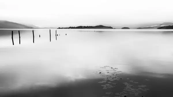 Lomond Gölü 'nün sakin tek renkli manzarası gökyüzünü yansıtan sakin bir göle sahiptir, tahta direkler ve uzak tepelerle..