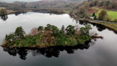 Huzurlu bir manzara, arka planda tepeler olan gölün sakin sularına yansıyan yemyeşil ağaçlarla kaplı küçük bir adayı sergiliyor..