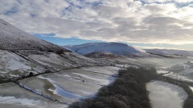 Bulutlu mavi gökyüzünün altında karla kaplı bir dağ manzarasının çarpıcı bir görüntüsü doğanın güzelliğini yakalıyor. Howgill Şelaleleri ve M6 otoyolunun üzerinde beyaz karla kaplı bir şekilde kış sabahının doğuşuna doğru uçuyor. Yorkshire Dale İngiltere.