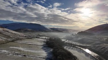 Sakin bir kış manzarası karla kaplı dağlar ve bulutlu bir gökyüzünün altında dolambaçlı bir nehir. Karla kaplı dağların, bir nehrin ve güneşin altında güzel bulutlu bir gökyüzünün bulunduğu çarpıcı bir manzara..