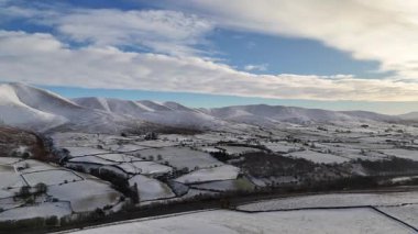 Bir hava manzarası, karla kaplı dağlık bir araziyi gösteriyor. Parçalı bulutlu bir gökyüzünün altından geçen bir otoyol. Karla kaplı bir dağ otoyolu bulutlu bir gökyüzünün altında kışın esen bir arazide esiyor..