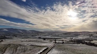 Bir hava manzarası, karların tepeleri örttüğü bir kış manzarası yakalar ve bir yol, parlak bir gökyüzünün altında gezinir..
