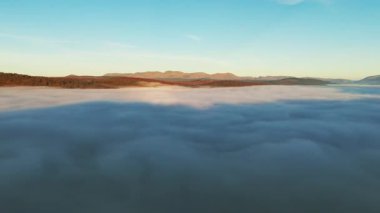 Şafak veya alacakaranlıkta berrak bir mavi gökyüzünün altında dağlarla kısmen gizlenmiş manzaralı bir yer. Parlak bir gökyüzünün altında kalın bir sisle kaplanmış bir dağ silsilesinin çarpıcı hava manzarası..