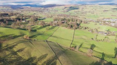 Uçan tepeler, tarlalar ve ağaçlarla dolu yemyeşil bir kırsal alan. Sahne güneş ışığıyla yıkanır, uzun gölgeler bırakır..