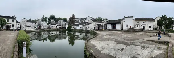 Geleneksel beyaz binaları olan antik bir Çin köyünün panoramik görüntüsü bulutlu bir gökyüzünün altındaki dingin bir göle yansıyor..