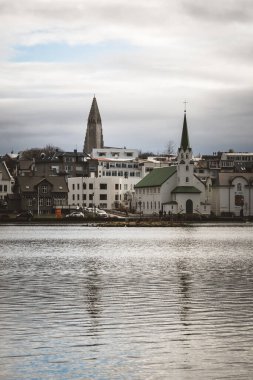 Gün doğumunda Reykjavk 'ta sakin bir göl, sabahın erken saatlerinde gökyüzünün renklerini ve bulutlarını yansıtıyor. Huzurlu ve atmosferik İskandinav kentsel manzarası.