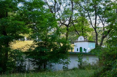 Kemerli yeşil kapıları ve grafitileri olan küçük beyaz bir yapı, ormandaki yoğun ağaçlar ve bitki örtüsüyle kısmen gizlenmiş..
