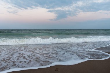 Yumuşak pastel gökyüzünün altında kumlu sahillere vuran okyanus dalgalarıyla sakin bir deniz manzarası, sakin bir akşam atmosferi ve kıyı güzelliğini yakalıyor..