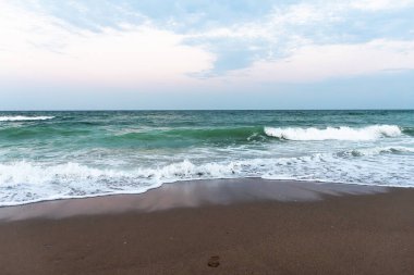Okyanus dalgaları kumlu sahilde kırılıyor pastel gökyüzü ve yumuşak bulutlar, fotokopi uzayı ile sakin deniz manzarası.