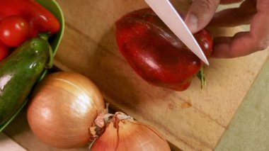 Erkek elleri, kırmızı dolma biberi, sebzelerle çevrili tahta kesme tahtasıyla keser. Çan biberi canlı renk ve doku gösteriyor. Sağlıklı ev yemeği kavramı.