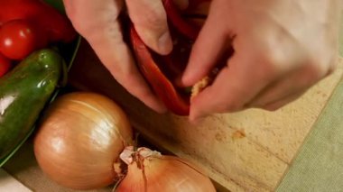 Erkek elleri ahşap kesme tahtasındaki kırmızı dolma biberi soğan ve domatesle temizler. Kırmızı dolma biber sebzelerle canlı kontrast gösteriyor. Yemek ve gıda tasarımında renk zıtlığı kavramı.