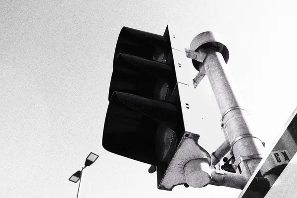 Trafik ışığının siyah ve beyaz bir fotoğrafı, kutuplarının açısal tasarımını ve kararmış lenslerini gösteriyor, kentsel manzaranın modern, endüstriyel estetiğini vurguluyor..