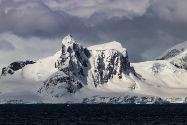 Tall mountain ridge on Antarctic Peninsula, topped by steep cliffs with rocks and blue ice. 