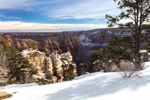 Grand Canyon view after recent snowstorm. Fresh white snow layered in foreground; Hoodoos and canyon wall dusted white in background. Blue sky and clouds above.