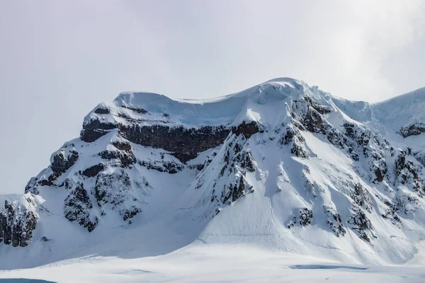 在南极半岛崎岖的山峰上 覆盖着积雪的陡峭的斜坡 下面的岩石暴露在外 蓝色的冰可见 背景是多云的天空 — 图库照片