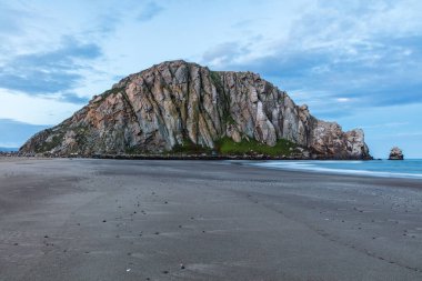 Şafak vakti ünlü Morro Rock Panorama, Morro Bay, California. Ön planda kumsal, sağda okyanus. Bulutlu mavi gökyüzü.