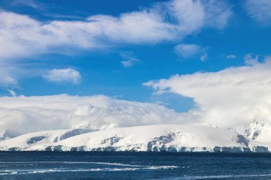 Antarktika yarımadasının kıyı şeridi derin karla kaplı. Önde okyanus, arkada mavi gökyüzü ve bulutlar. 