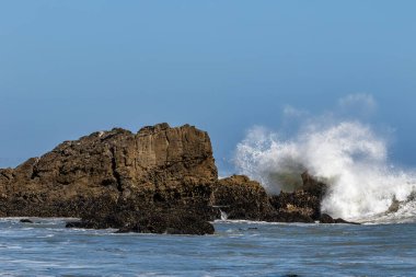 Malibu, California açıklarındaki büyük kayalara karşı dev dalgalar kırılıyor. Sprey havada yüksekte uçuyor, mavi gökyüzü uzakta.. 
