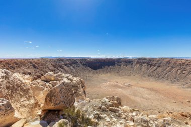Winslow, Arizona yakınlarındaki Barringer Meteor Krateri 'nde gözle görülür bir gözlem platformu var. Eskimiş kayalar, ön planda çöl bitkileri. Gökyüzü açık mavi. 