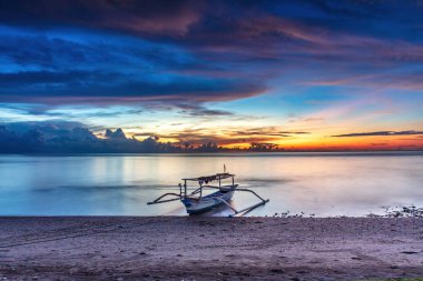 Okyanus batımı Lovina Sahili, kuzey Bali, Endonezya. Ön planda teknesi olan plaj; güneş renkli bulutlarla çevrili batıyor. Güneşin sudaki yansıması. 