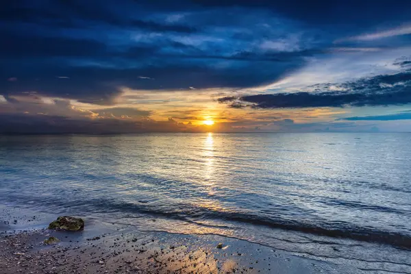 Okyanus batımı Lovina Sahili, kuzey Bali, Endonezya. Sahilde ön planda; güneş renkli bulutlarla çevrili batıyor. Güneşin sudaki yansıması. 