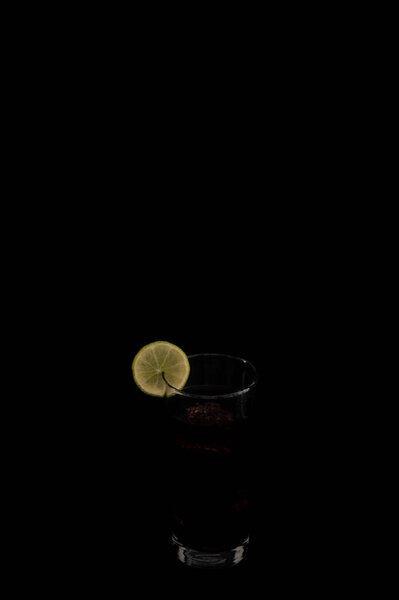 A glass of dark beverage with a slice of lime on the rim, set against a black background