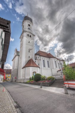 Nesselwang, Germany - July 06, 2023: Extreme wide angle view of the Saint Andreas Church with bell tower in Nesselwang from the north against a cloudy sky. clipart