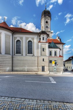 Nesselwang, Germany - July 06, 2023: South facade of the Saint Andreas Church with signposts and a view over the main street against a blue sky with clouds. clipart
