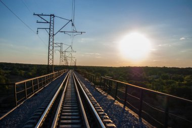 Ağustos güneşinin batışında tek raylı bir demiryolu köprüsü. Pistin kenarlarında güvenlik cepleri olan yaya yolları var. Solda bir elektrik hattı var..