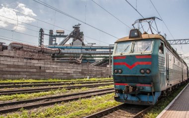 İstasyona kırmızı çizgili mavi bir yolcu lokomotifi geliyor. Arka planda fabrika binaları var..