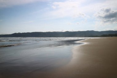 Sakin, yansıtıcı su alçak gelgitte sahil şeridinde. Okyanus, gökyüzü ve uzak tepe çizgisi.