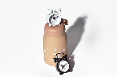 A creative still life featuring a white and black mini alarm clock placed with a beige water bottle on a white background, casting shadows.