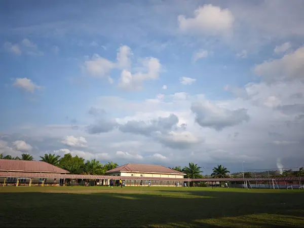 Güzel mavi gökyüzü ve arka planda dağınık beyaz bulutlar olan yeşil bir okul alanının geniş bir görüntüsü.