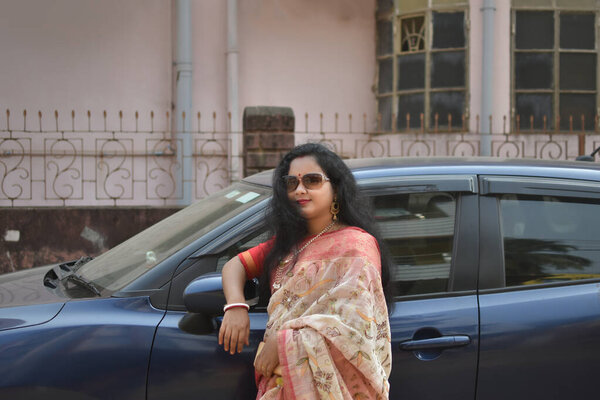 Молодая счастливая, красивая индийская женщина или домохозяйка, стоящая рядом со своей серой машиной на дороге с кошельком в руке.