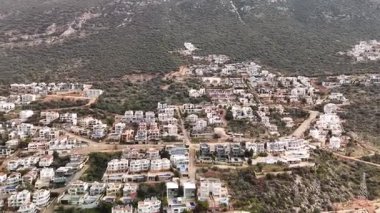 Kalkan, Türkiye 'nin havadan 4K görüntüsü. İnsansız hava aracı dağın manzarasını yakalıyor ve tepe boyunca yeşil ve deniz manzaralı zarif beyaz villalar inşa ediliyor.