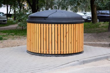 Şık bir ahşap tasarımı olan şehir çöp tenekesi asfalt bir yüzey üzerinde duruyor ve şehrin atık yönetimi altyapısı, açık hava manzarasına doğal bir zarafet katıyor.